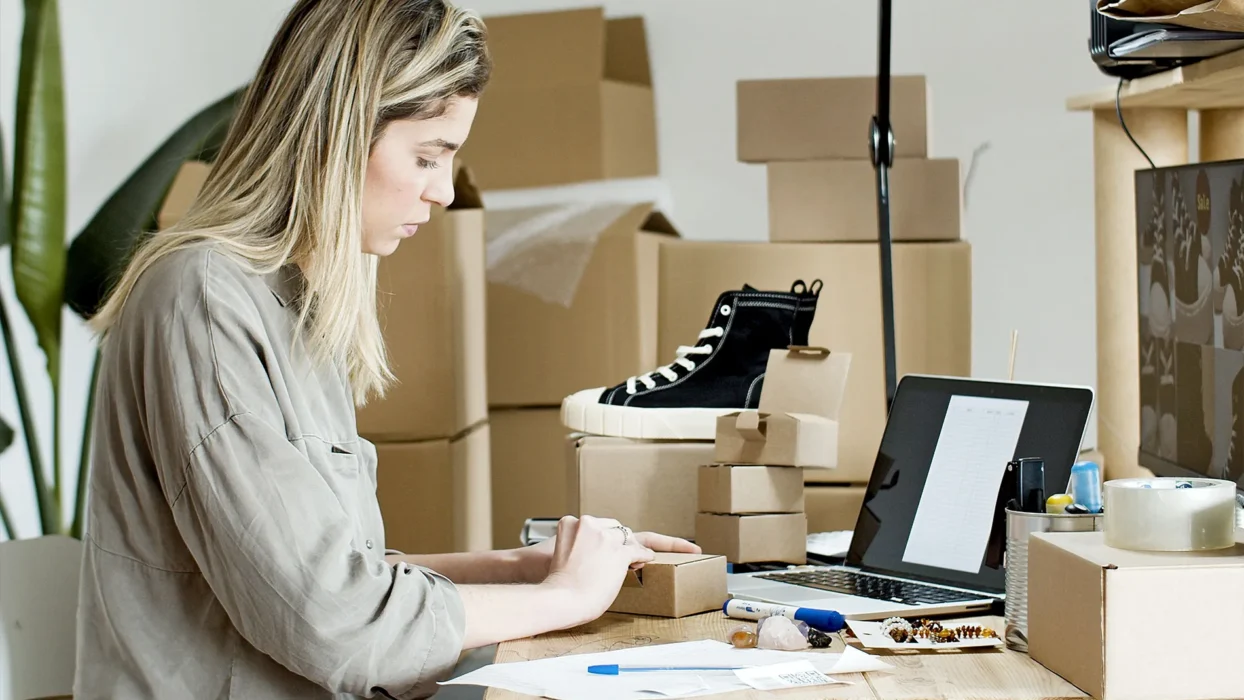 A woman packing boxes for her online store, surrounded by packaging materials and a laptop
