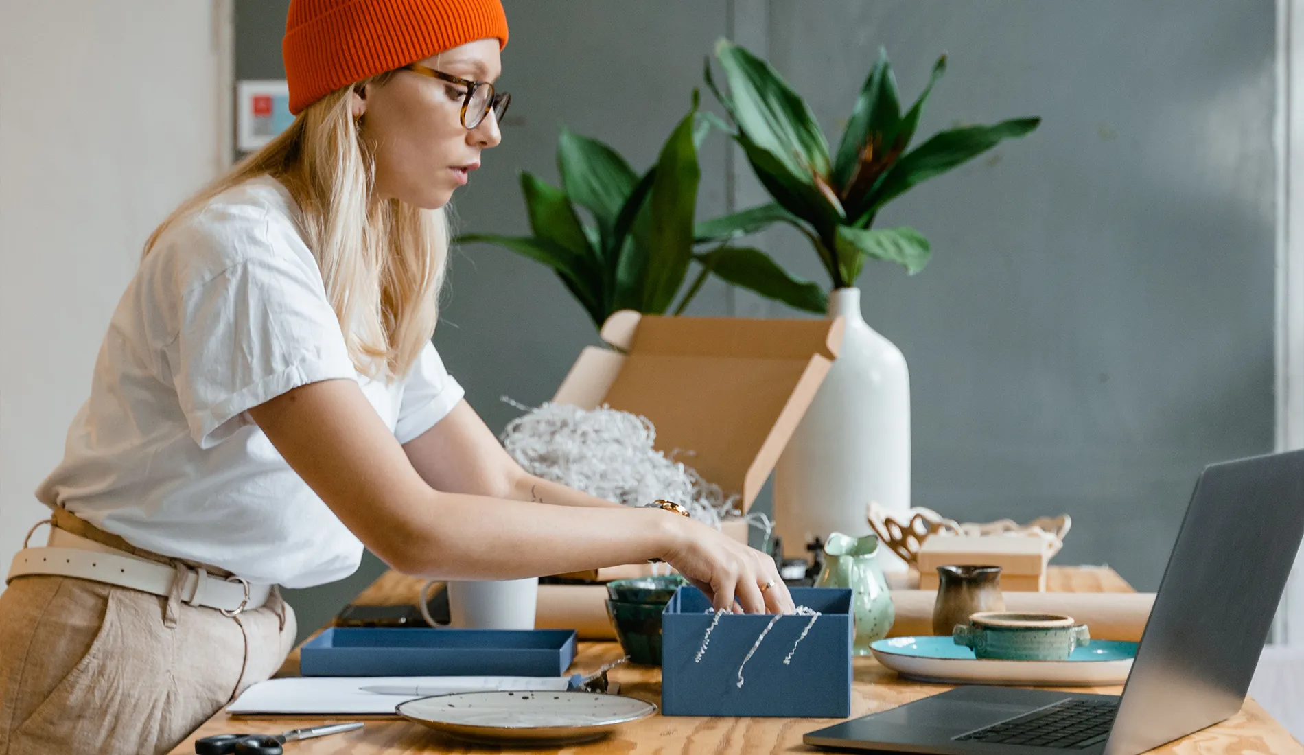 A young woman in a beanie packs boxes at a stylish wooden table in a home office